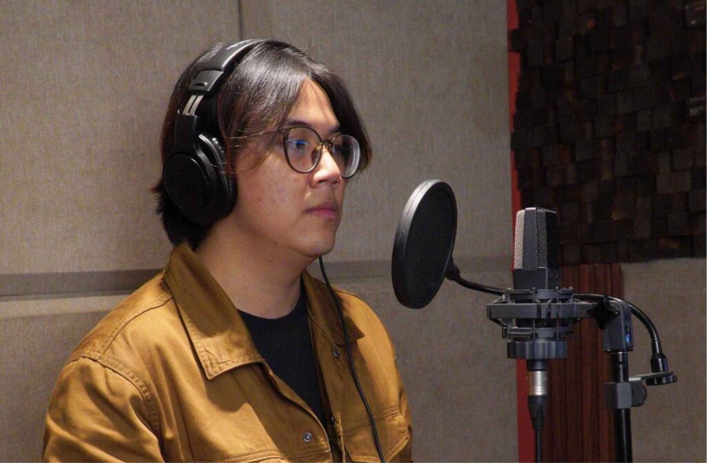 Voice actor recording in a professional studio with headphones and a condenser microphone.